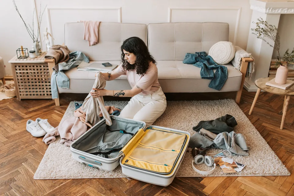Woman with curly hair sitting on a rug in front of a sofa, packing clothes into an open suitcase