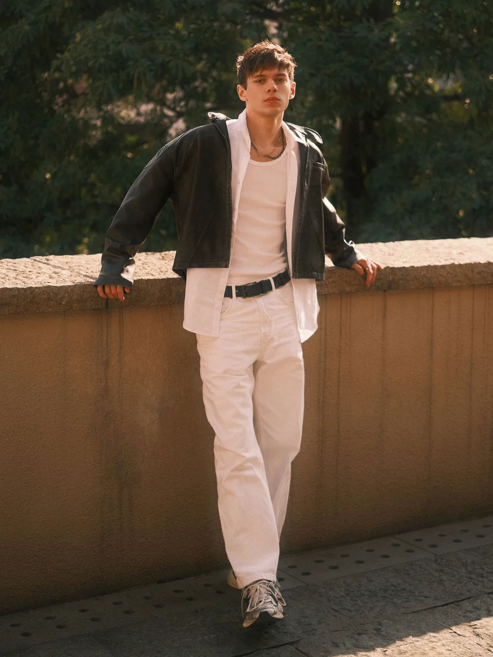 Man in a black leather jacket, white shirt and white trousers leaning on a wall