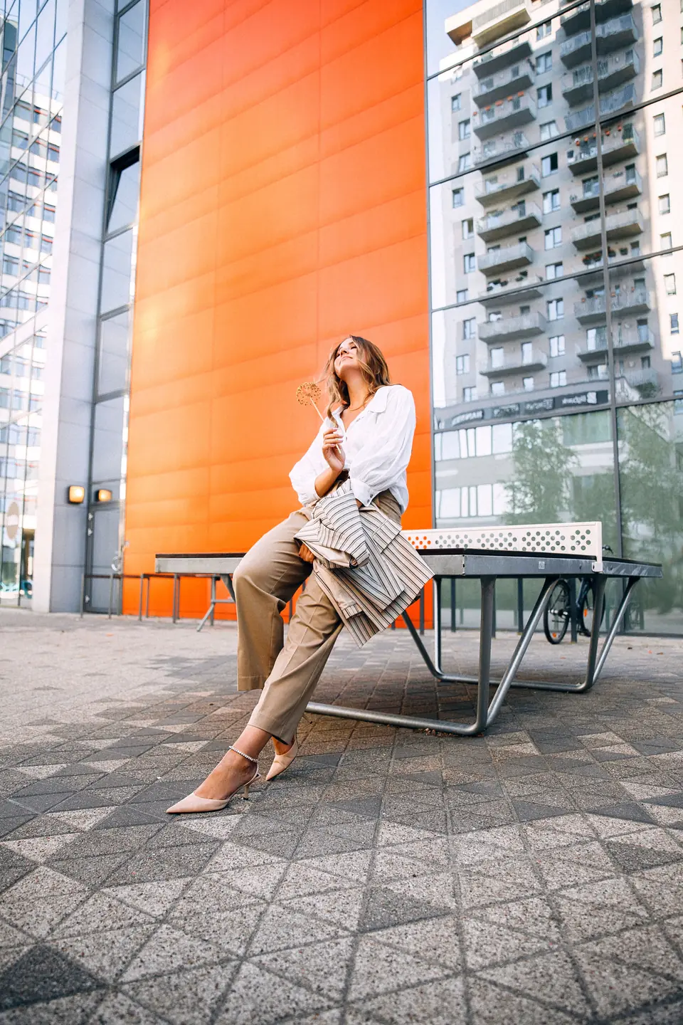 Woman in a white blouse and beige trousers sitting on a table-tennis table in front of an orange facade, a striped blazer resting on her knee