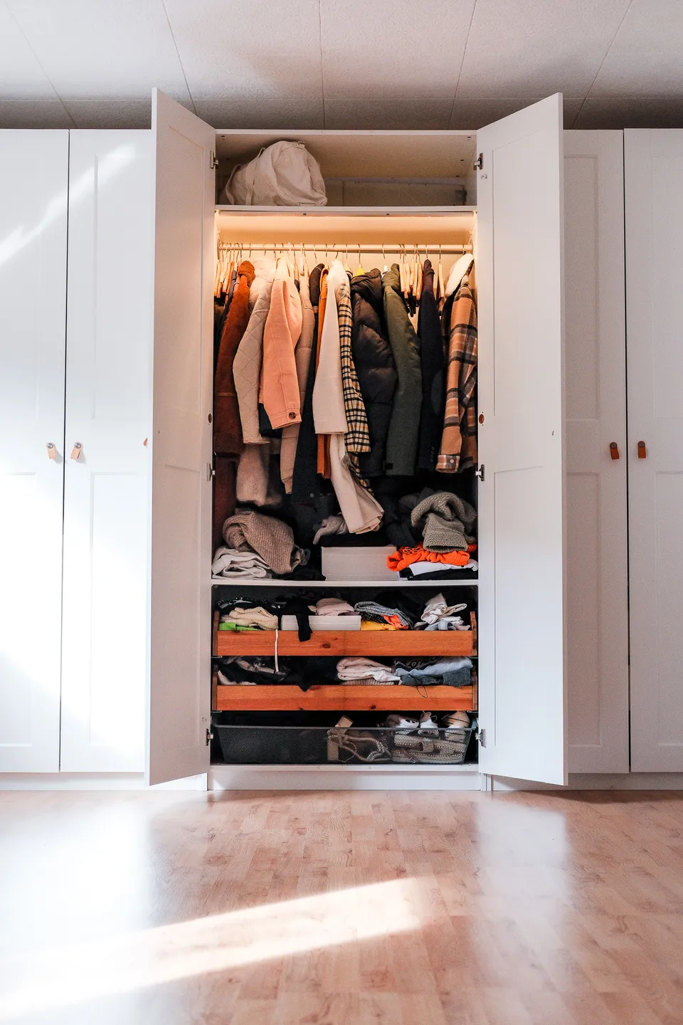 Open wardrobe with jackets on wooden hangers, folded sweaters and two wooden drawers below