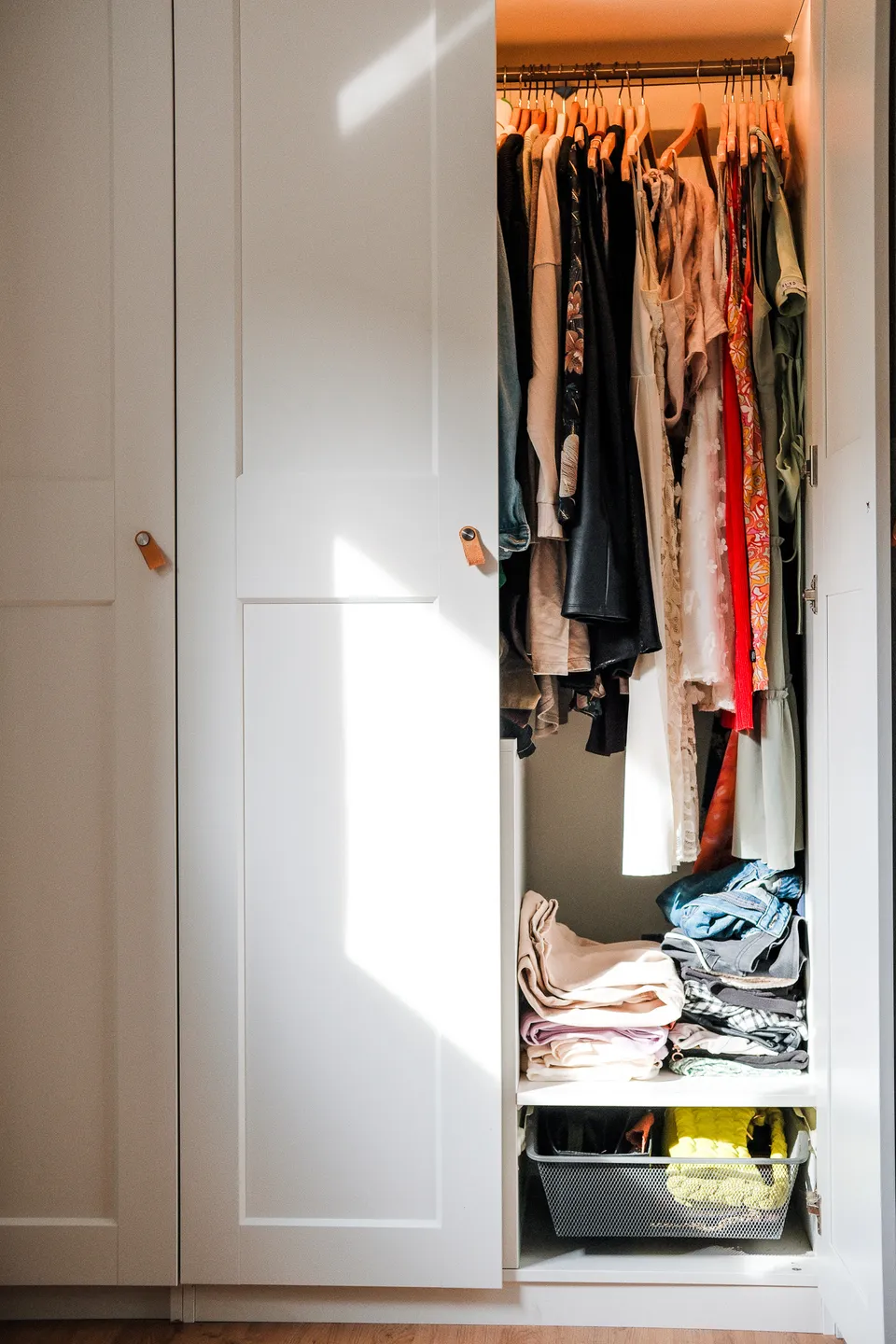 Wardrobe with one door open: dresses and blouses on hangers, folded jeans and a basket below